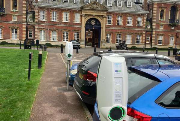 2 EV chargers next to row of parked cars with college building in the background